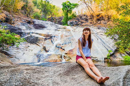 Asian woman travelers are smiling and relaxes sitting on the stone in front of the waterfall at Namtok Kaew Chan\n, Ratchaburi, Thailand.