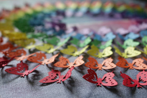 Stock photo of close-up of gay themed homemade crafting art work, using rainbow butterfly punch with cut out card and paper butterflies, embossed and glued on abstract watercolour painting that has been painted on a stretched canvas. This original craft art work of rainbow coloured made of multicoloured cut out butterfly shapes (Monarch paper butterflies - purple, blue, green, orange, yellow and red colours) is like the rainbow LGBTQI gay flag, with the colourful arts and crafts project displayed on wall as background wallpaper. imagen de primer plano de la obra de arte artesanal casero de temática gay, utilizando el ponche de mariposa arco iris con tarjeta cortada y mariposas de papel, en relieve y pegado en pintura abstracta de acuarela pintada sobre un lienzo estirado. artesan