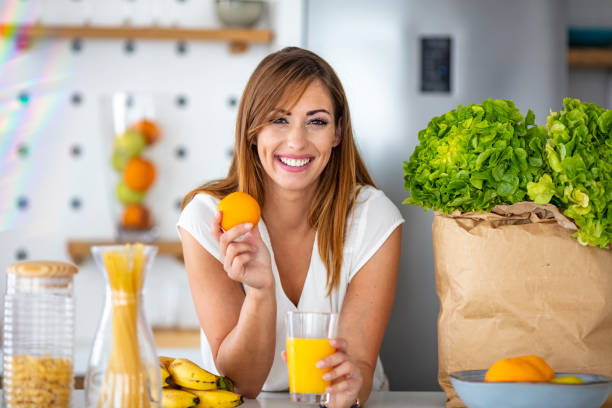 Pretty woman holding glass with tasty juice Young joyful woman drinking orange juice and standing near a kitchen table. Close up of a woman drinking juice in her kitchen. Fit smiling young woman preparing healthy fruit juice vitamin c stock pictures, royalty-free photos & images