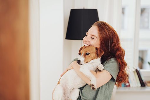 Happy young woman cuddling her little dog in her arms as she stands indoors in her living room