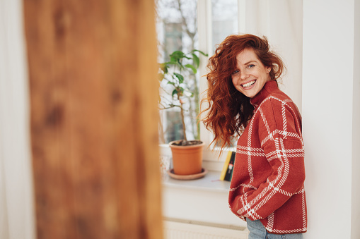 Friendly young redhead woman relaxing at home in a warm winter sweater with her hands in her pockets grinning at camera