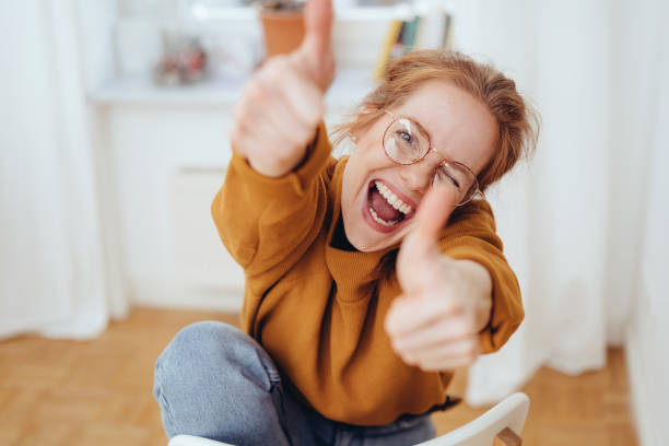 Happy girl showing thumbs up gestures Happy pretty girl in glasses showing thumbs up gestures, sitting on chair in the room. Close-up portrait from high angle joy stock pictures, royalty-free photos & images