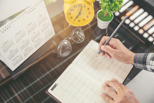 Woman's hand planner or organizer writing daily appointment, mark, and noted schedule (meeting) on timetable or diary at office desk.Calendar, agenda, book, glasses, and clock on table.Planner concept.