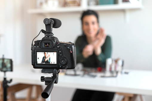 Young Woman Vlogging Herself About Beauty Products. Woman Making a Video for Her Blog on Cosmetics Using a Tripod Mounted Digital Camera. Young Female Blogger on Camera Screen Holding Cosmetics.