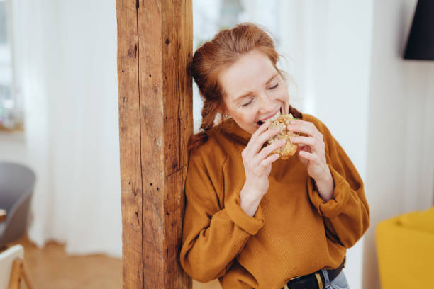 Hungry young woman taking a bite Hungry young woman taking a bite of a healthy wholewheat sandwich with a happy smile leaning on a wooden pillar indoors at home sandwich healthy lifestyle healthy eating bread stock pictures, royalty-free photos & images