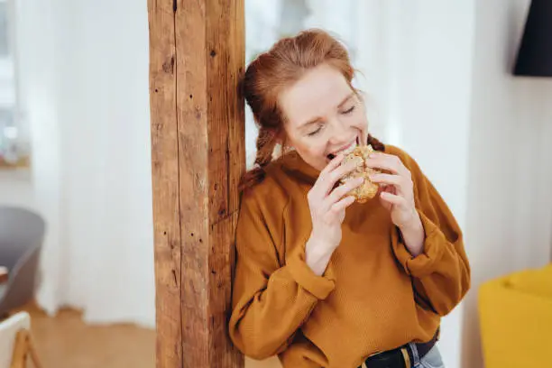 Hungry young woman taking a bite of a healthy wholewheat sandwich with a happy smile leaning on a wooden pillar indoors at home