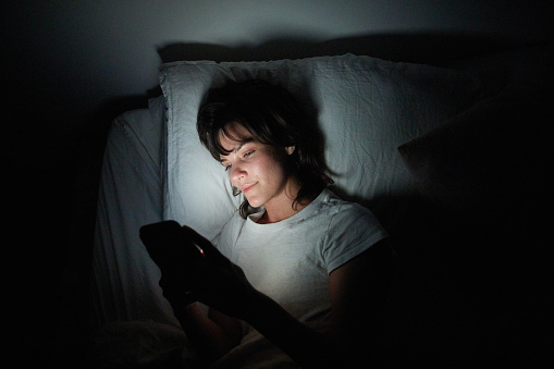 Young woman lying in bed at night with her face illuminated by the display of her cellphone