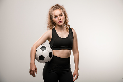 Pretty young female soccer player with long blond curly hair holding ball between arm and waist while standing in front of camera