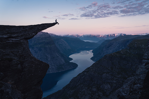 A woman sitting on the edge of Trolltunga in a beautiful morning sunrise in summer season, Odda town, Norway, Scandinavia, Europe