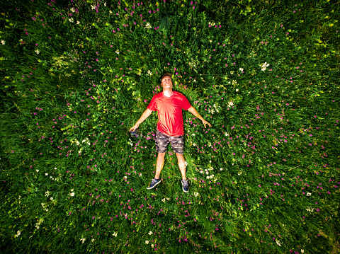 Drone shot depicting a high angle view of man lying down in a field of yellow flowers. The camera is pointing directly down at the man, who is wearing casual t shirt, shorts and baseball cap, who is surrounded by flowers and lush green vegetation. Room for copy spac.