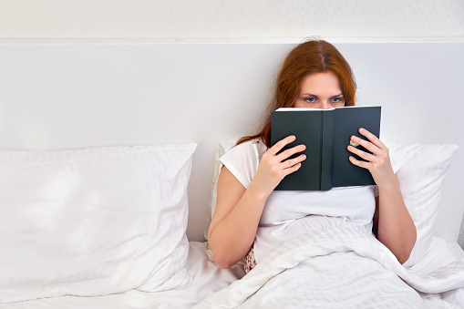 A single woman reads a book lying alone in a double bed. Family relationships in isolation due to coronavirus.