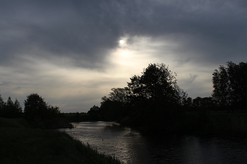 Evening landscape. Dark time. Black trees against a gray sky with clouds on the Bank of a small river.