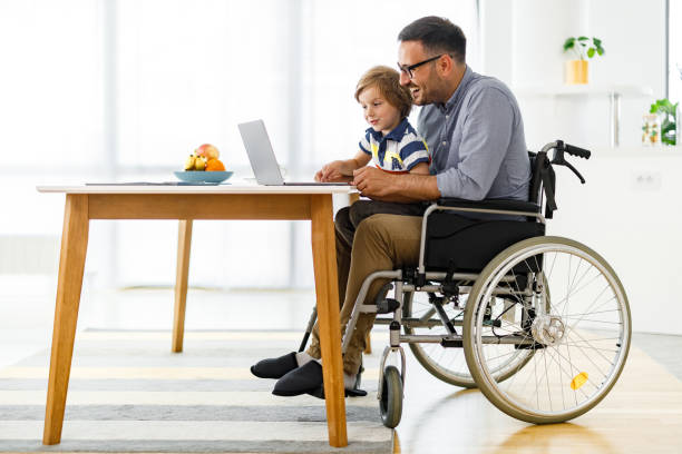 Happy disabled father and his small boy using computer at home. Happy single father in a wheelchair and his small son using laptop at home. Copy space. accessibility for persons with disabilities photos stock pictures, royalty-free photos & images