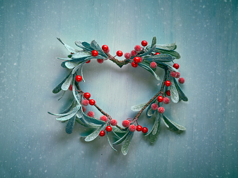 Decorative Christmas heart shaped wreath with frosted mistletoe leaves and red berries hanging on a light textured door. Toned Xmas decoration background in green and red.