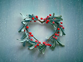 Decorative Christmas heart shaped wreath with frosted mistletoe leaves and red berries hanging on a light textured wooden door