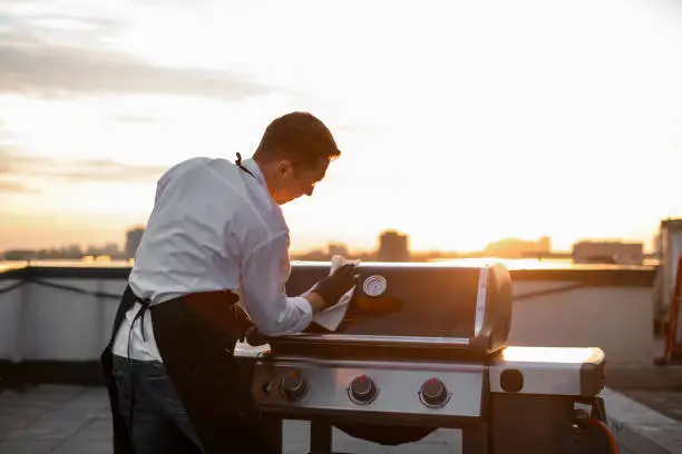 cook man in black gloves and aprons wipes the grill equipment for barbecue. Sunny sunset