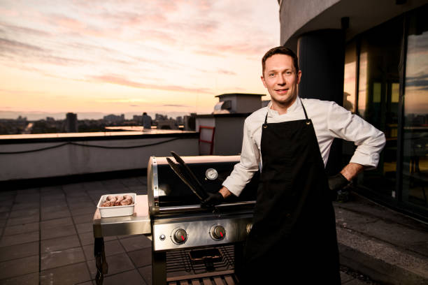 笑顔の男性シェフは、彼の手にトングを保持し、バーベキュー機器の近くに立っています。 - barbecue grill chef barbecue sausage ストックフォトと画像