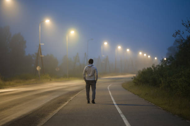 un giovane cammina lentamente sul lungo marciapiede sotto i lampioni nella notte d'estate. passare del tempo da solo. vista posteriore. - walking at night foto e immagini stock