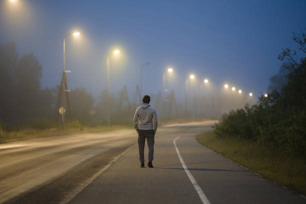 One young man slowly walking on long sidewalk under street lights in summer night. Spending time alone. Back view.