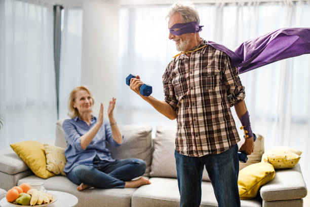 I'm a strong superhero! Happy mature man having fun while exercising with weights at home and pretending to be a superhero. His wife is supporting him in the background. senior bodybuilders stock pictures, royalty-free photos & images