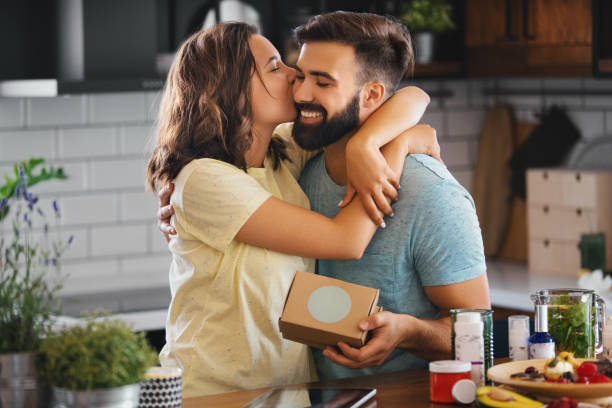 A surprise gift for my love. Young couple in the kitchen. The man surprised his girlfriend with an unexpected gift. She is happy. Hugging him and kissing. unusual couple stock pictures, royalty-free photos & images