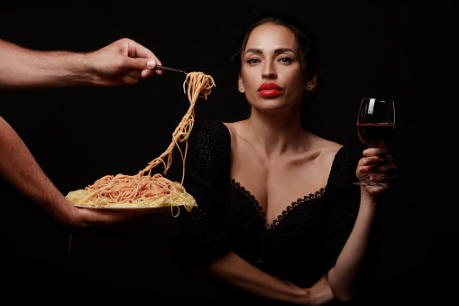 A beautiful woman eats spaghetti and drinks wine. A fork with a plate is held by a man's hand. On a black background. Italian cuisine.
