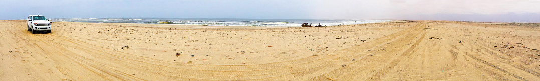 Waters edge, Skeleton Coast National Park, Namibia, Africa. Panoramic wide angle view of coastline of Skeleton Coast with a solitary vehicle driving on the treacherous sands along this notorious coastline famous for wrecking ships due to the dangerous Benguela current and fog