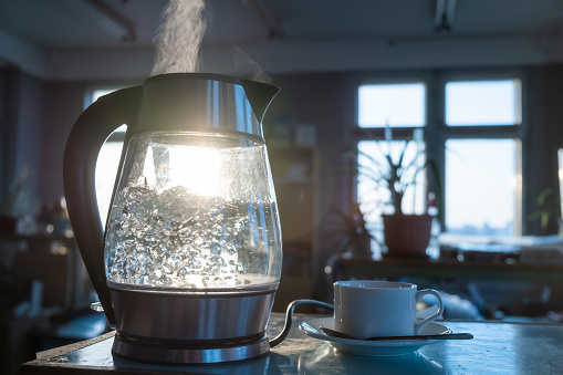 A transparent kettle of water boils against the background of the sunset shining through the window. The concept of coffee break and end of the working day.