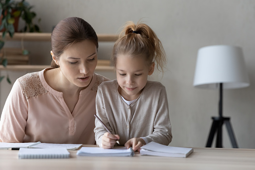 Loving young Caucasian mother sit at desk prepare homework, handwrite in notebook with small daughter, caring mom or nanny studying learning with little girl child, do homework, education concept