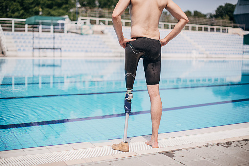 One man, disabled young man with one prosthetic leg, standing alone by the swimming pool outdoors.