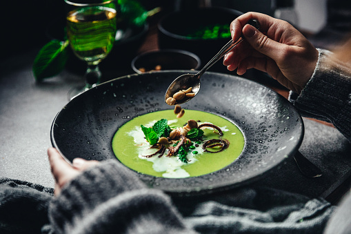Close-up of woman garnishing green peas and cream soup. Female hand adding roasted garlic to vegan soup on table.