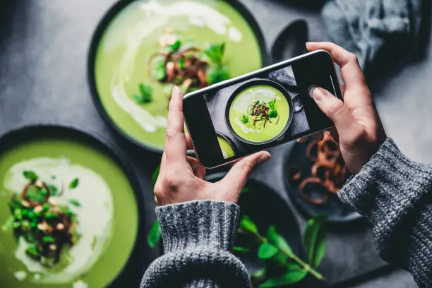 Close-up of a woman taking a photo of green cream soup with her mobile phone. Female hand photographing green soup.
