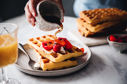 Close-up of a female hand pouring maple syrup on waffle. Woman serving sweet and tasty breakfast.