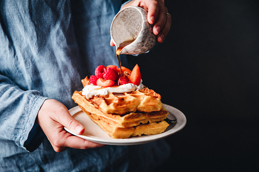 Close-up of a female hands holding a plate with waffles Greek yogurt and strawberries. Woman pouring maple syrup over waffle in a plate.