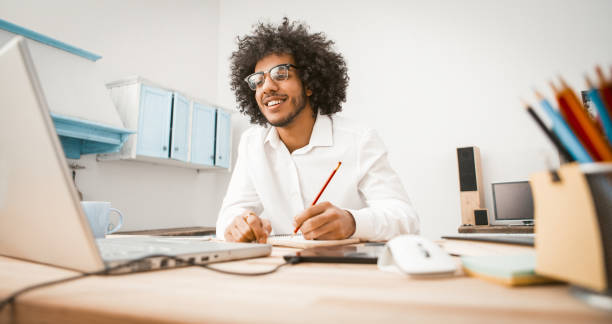 istock Happy millennial work at home workplace. Shaggy young man takes notes while looking at a computer monitor sitting at table studio apartment. Distance learning or home education concept. Toned image 1249209711