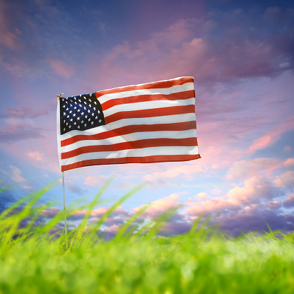 Conceptual image of waving American flag at flagpole on green landscape over cloudy dusk time sky