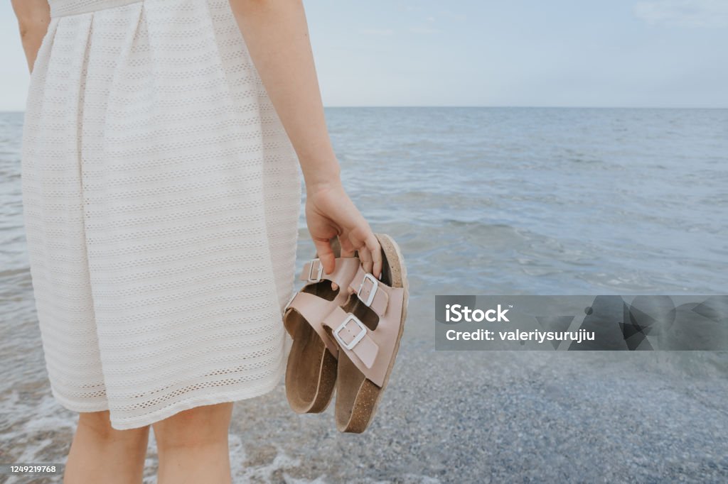 woman with beach slippers woman in a white dress holds beach slippers in her hands and walks along the shore of the blue sea Women Stock Photo