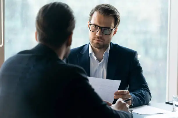 Serious attentive HR manager wearing glasses looking at job applicant, holding cv, listening to candidate on interview, strict employer, recruiter making decision about hiring, difficult negotiations
