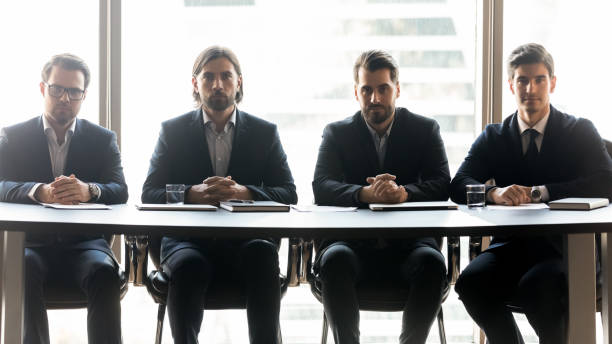 quatre hommes d’affaires sérieux s’asseyant dans la rangée à la réunion - exam business caucasian board room photos et images de collection