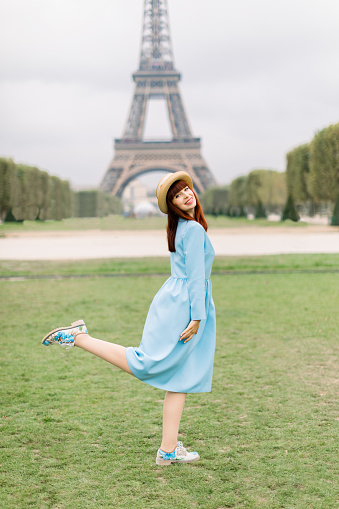 Portrait of a young excited smiling woman in straw hat and blue dress posing with one leg up,standing on green grass in front of the Eiffel tower in Paris.