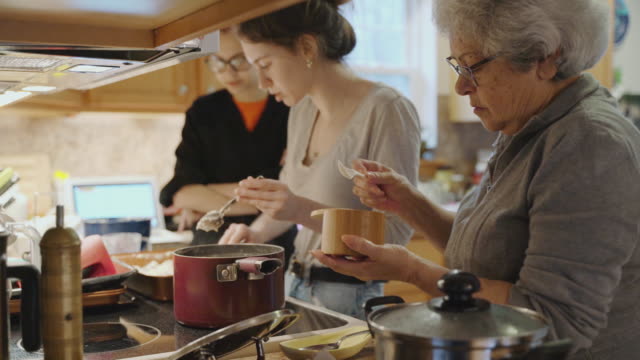 Grandmother is adding salt into the pot she is using to cook with at the stove while her two granddaughter is also are cooking in the background.
