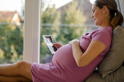 Side view, close up of a pregnant woman in comfortable lavender color maternity dress, sitting next to a window in day time and looking at the ultrasound picture of her baby.