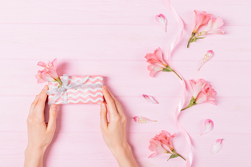 Top view of female hands holding gift box next to composition of fresh flowers, petals and silk ribbon on pink background.