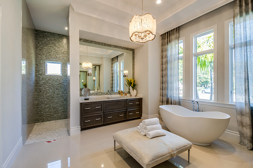 Freestanding tub and ottoman stool in center of bathroom with shower in the background
