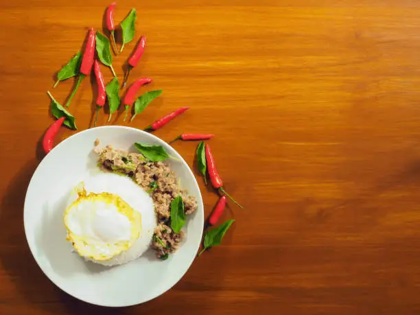 A white dish with steamed rice overlayed with fried eggs and stir-fried with basil with pork Beside, there are chilli and freckles because they are beautifully decorated. The top view on the background is hardwood.