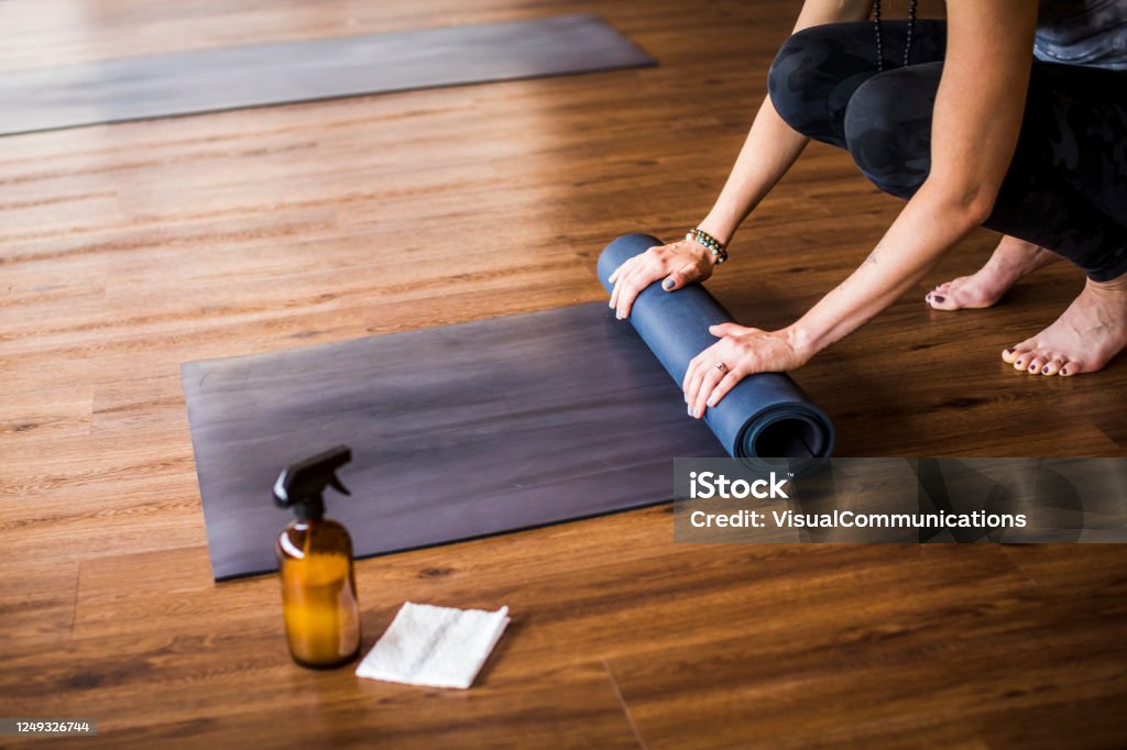 Woman rolling up yoga mat with sanitizer nearby. Yoga training concept during covid-19 in indoor studio. Cleaning Stock Photo