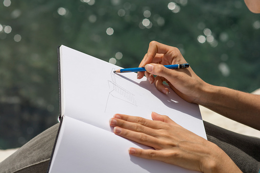 Woman hands drawing a sketch on a white page of a sketchbook, using a blue 6B pencil, Outdoors