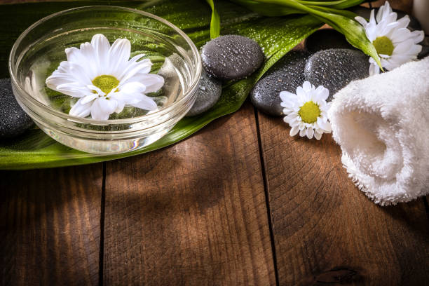 High angle view of some wet hot stones for a spa therapy, a crystal bowl full of water with a white flower and a white towel on a wooden table. Predominant colors are white, brown and green. Objects are at the top of the image leaving a useful copy space at the lower side on the table. Studio shot taken with Canon EOS 6D Mark II and Canon EF 24-105 mm f/4L