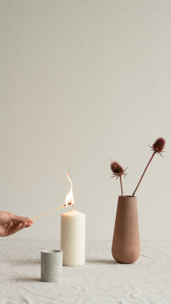 Young female hand burning aromatic candle between vase with plant and glass Young female hand burning white aromatic candle standing between small ceramic glass and brown clay vase with wildflowers on table hygge photos stock pictures, royalty-free photos & images