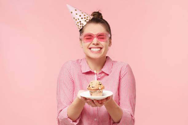 Excited funny teenage girl wearing colored glasses and birthday hat, holding cupcake on plate with two hands, making wish with closed eyes, isolated on pink background Excited funny teenage girl wearing colored glasses and birthday hat, holding cupcake on plate with two hands, making wish with closed eyes, isolated on pink background woman birthday cake stock pictures, royalty-free photos & images